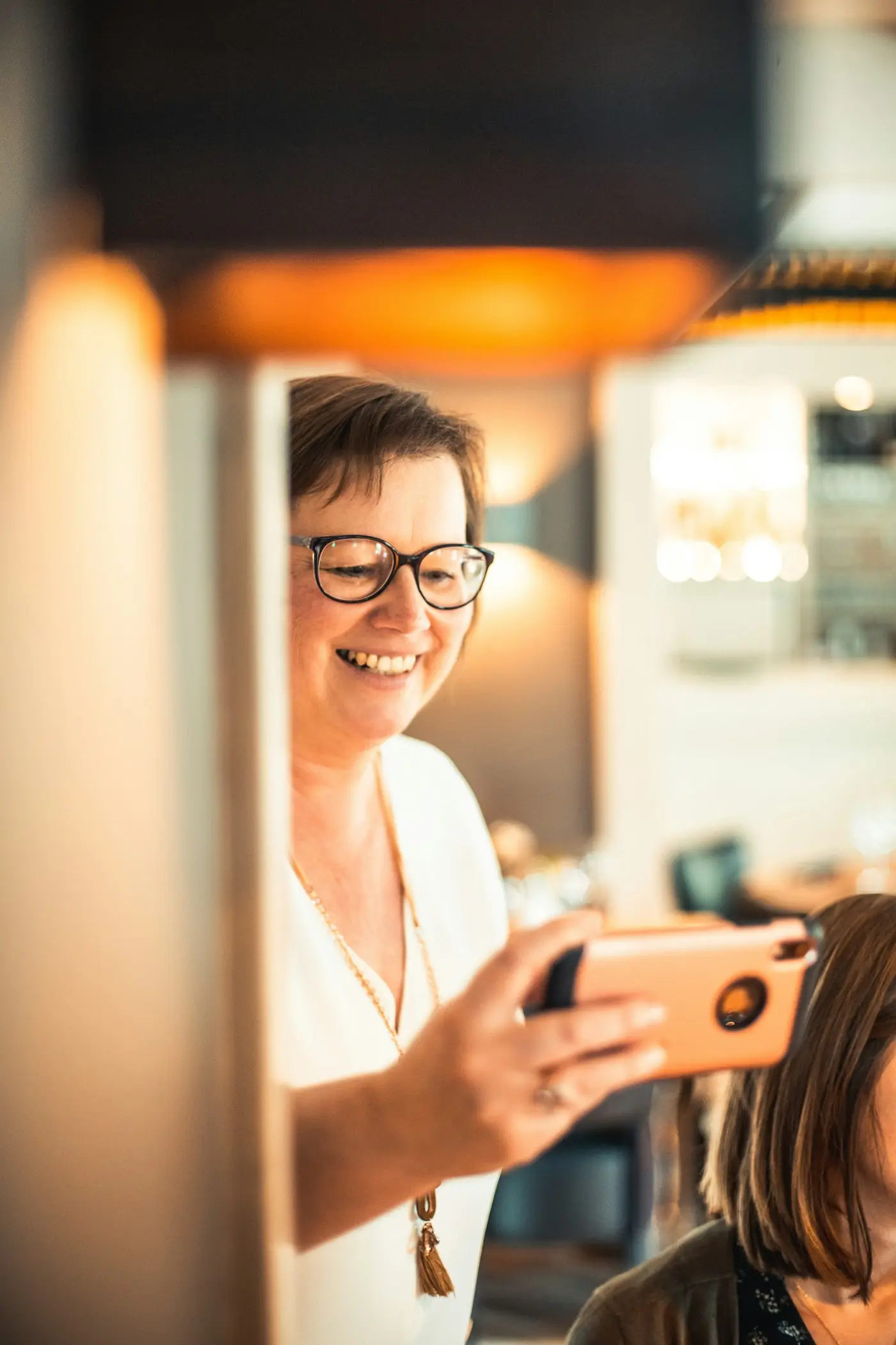 Profissional de óculos sorrindo enquanto olha o celular no salão, sob luz quente